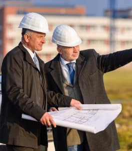 Two men in suits, ties, and hard hats holding blueprints on a work site