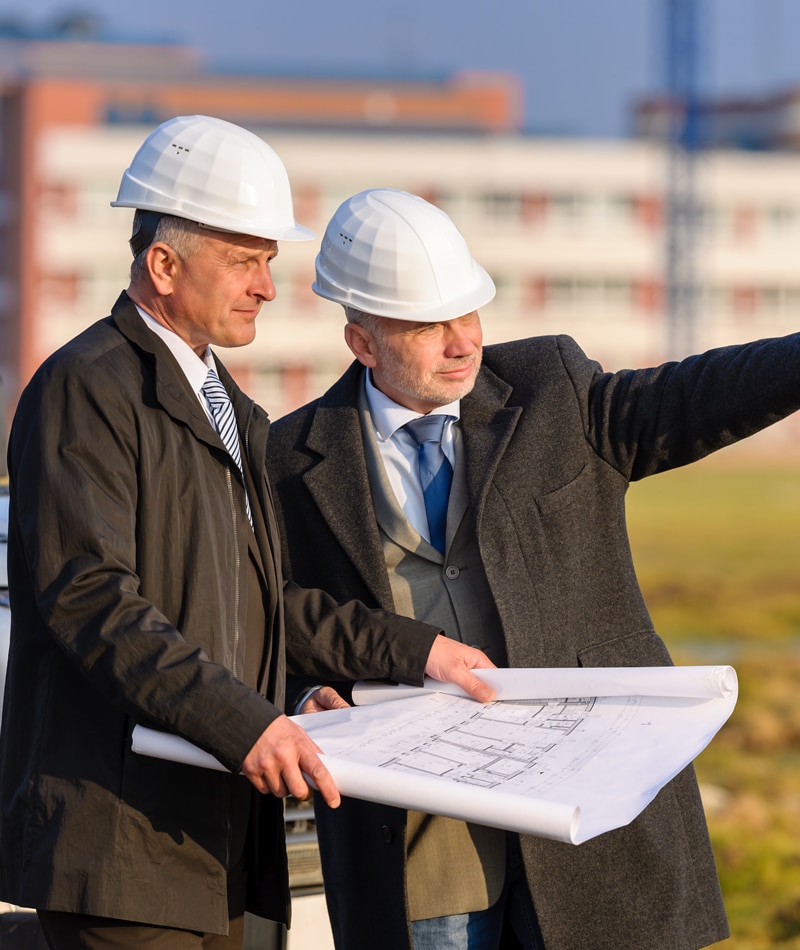 Men wearing suits, ties, and hard hats on a work site with blueprints
