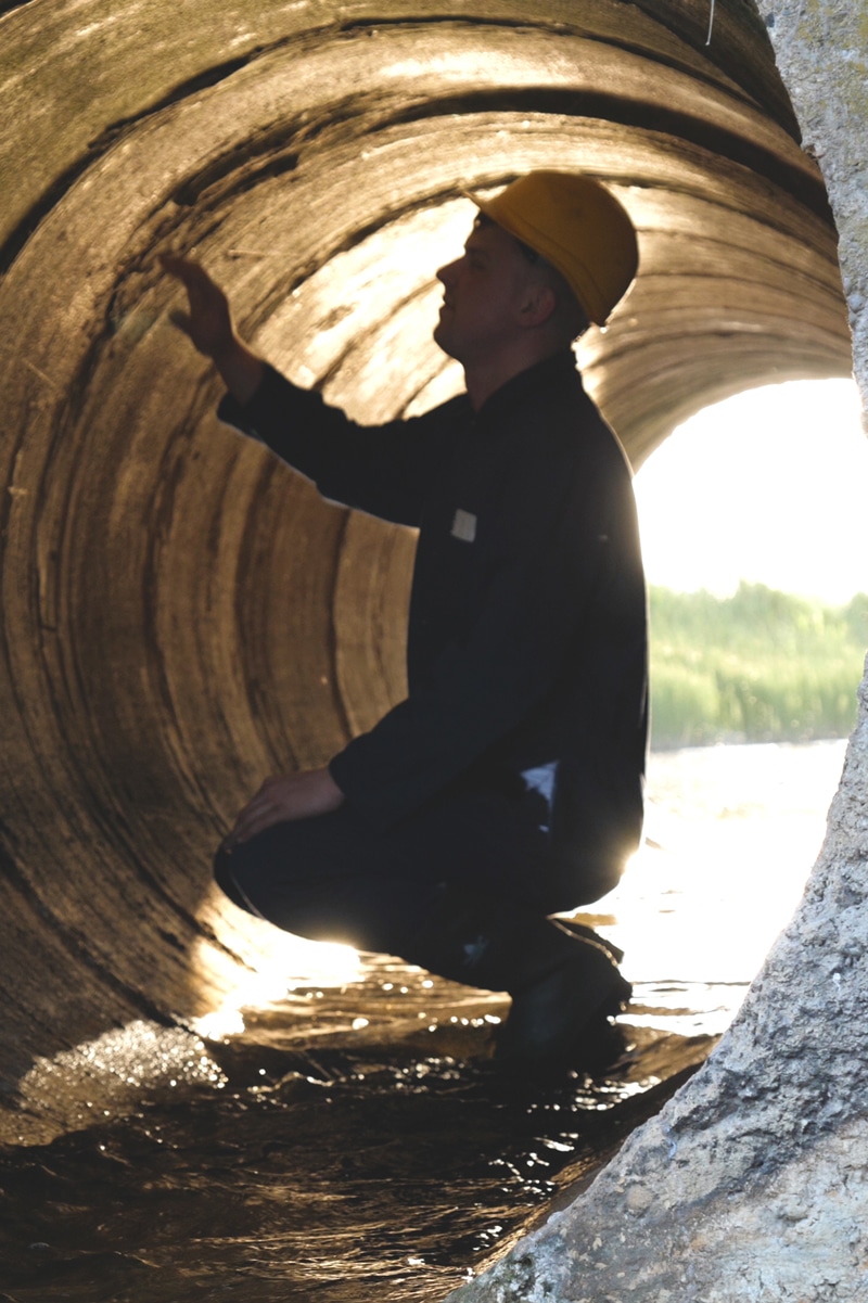 A man in a hard hat squatting inside of a large drainage pipe to inspect it
