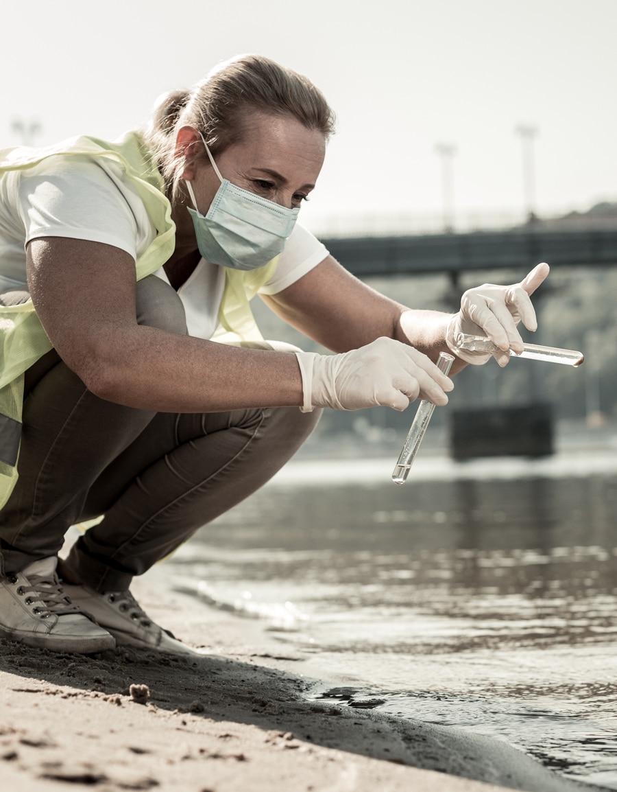 Woman in a mask and reflective vest holding two test tubes of water samples, pouring one tube into the other as a part of the Impaired Waters and TMDL Program
